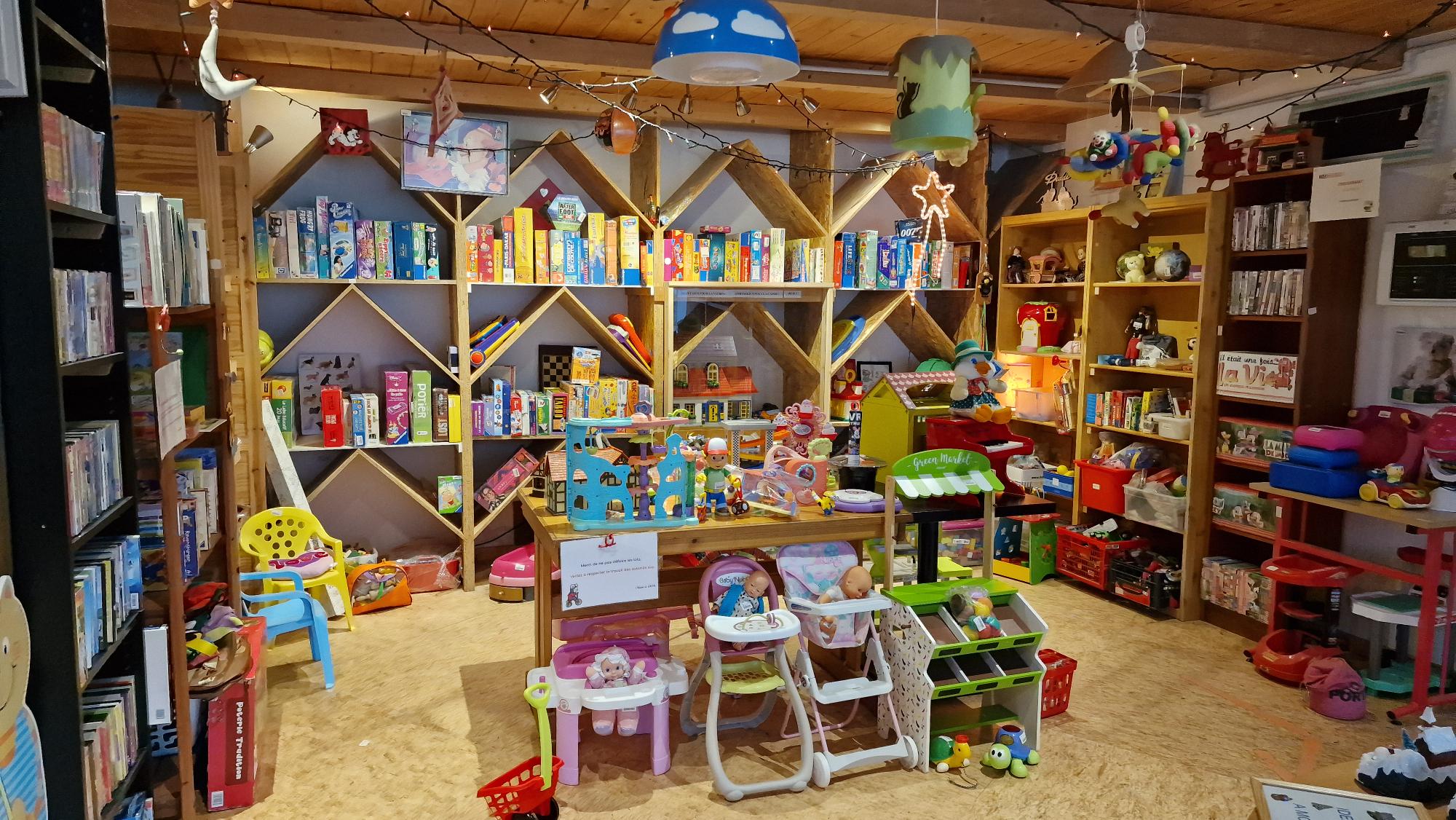 Une table centrale avec des chaises de bébé et jouets, entourée d'étagères remplies de livres et jeux pour enfants, dans une pièce en bois éclairée par des guirlandes.