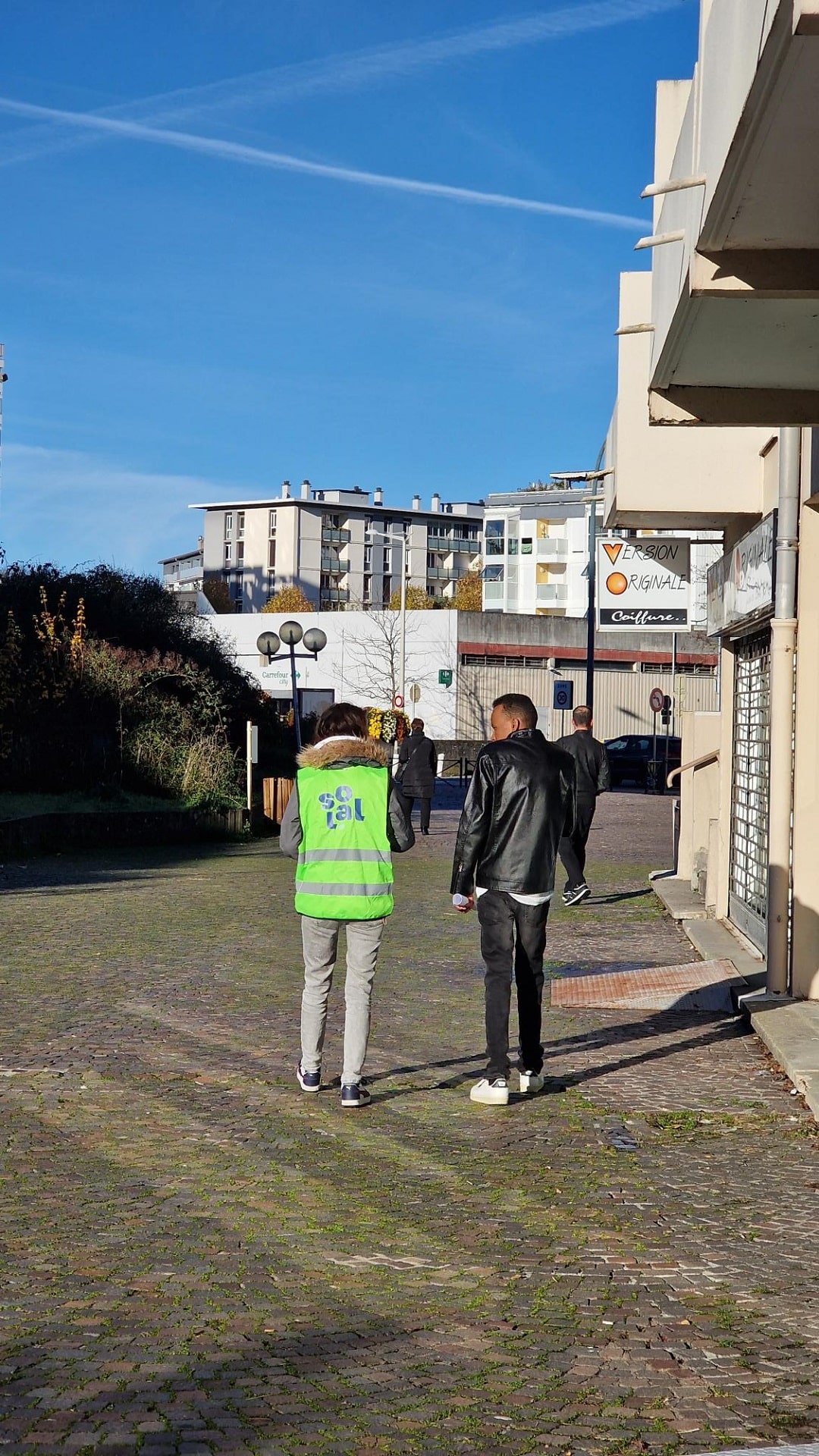 Deux personnes, l'une en gilet fluorescent, l'autre en veste cuir, marchent sur un chemin pavé bordé de bâtiments modernes sous un ciel bleu.